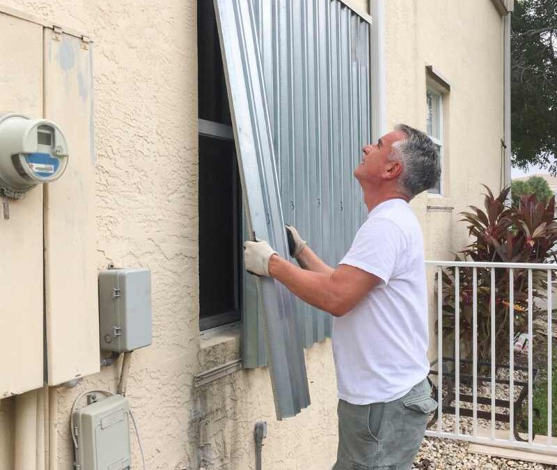 A man in a white t-shirt installs a metal storm shutter on a beige house